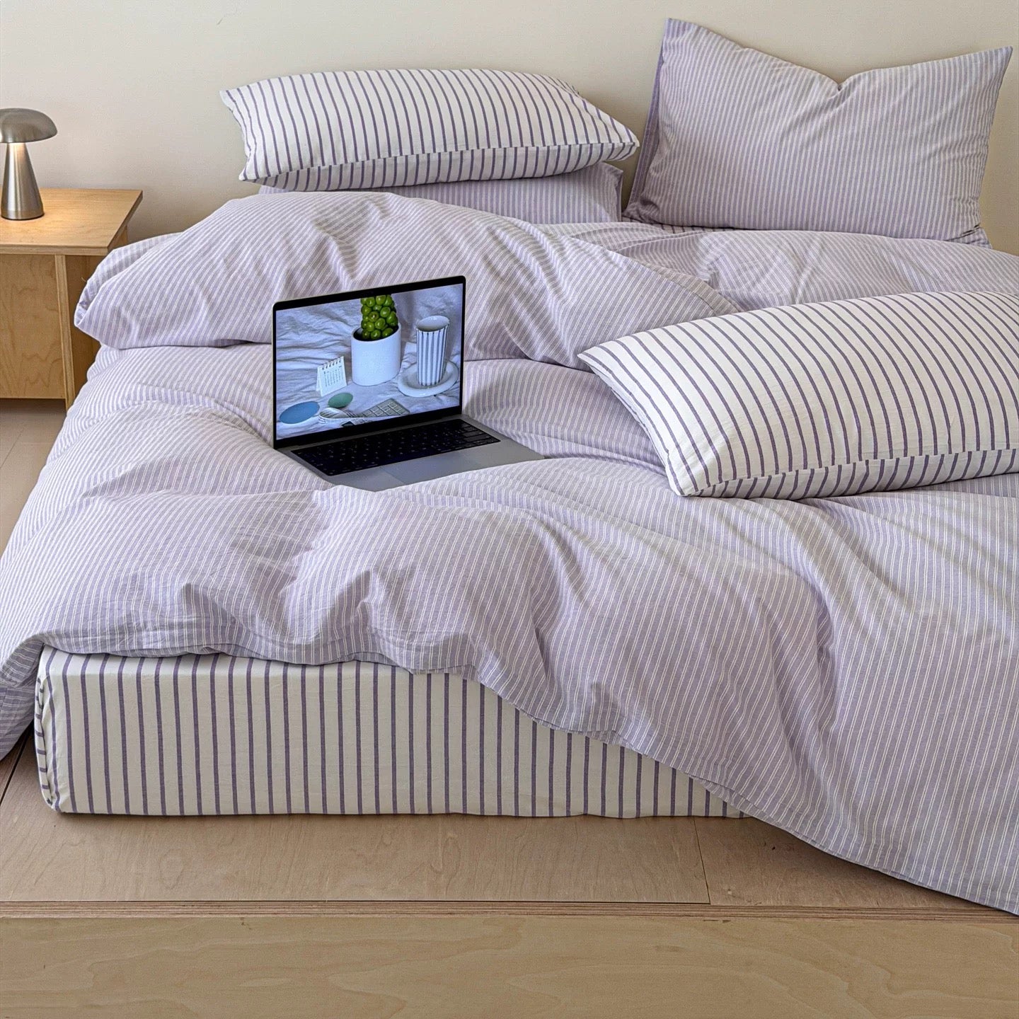 A bed dressed in light lavender and white striped bedding with matching pillows, shown in a bedroom setting with a laptop on the duvet.