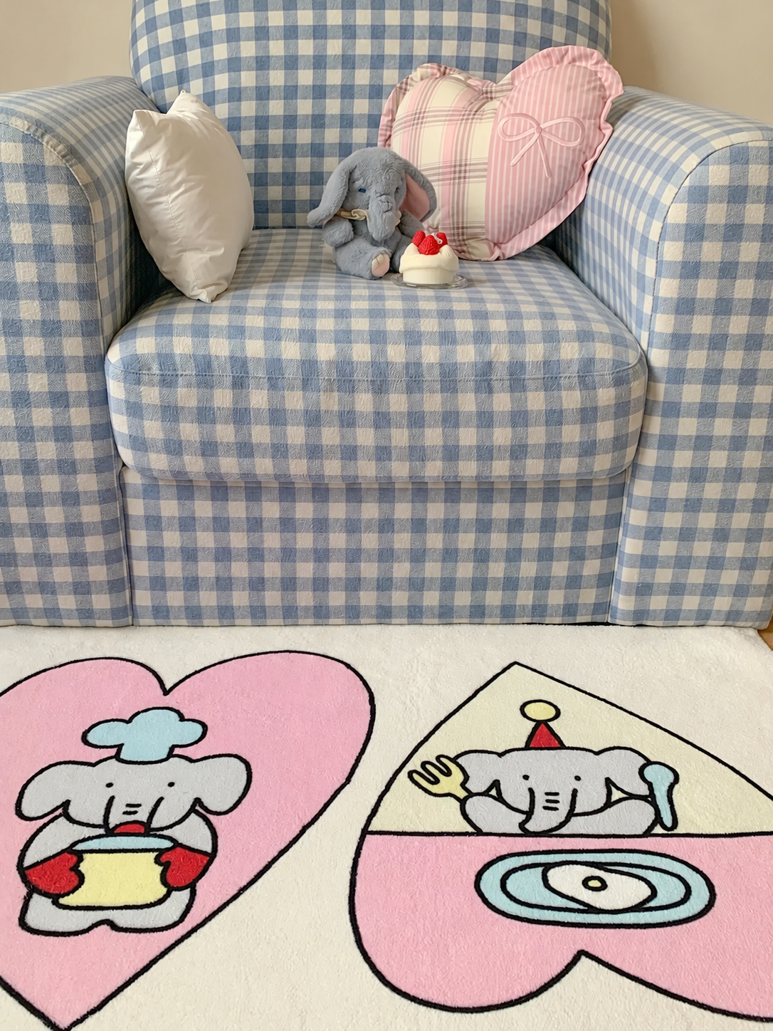A Kawaii rug with pink heart panels and little elephant characters sits in front of a blue checked armchair.