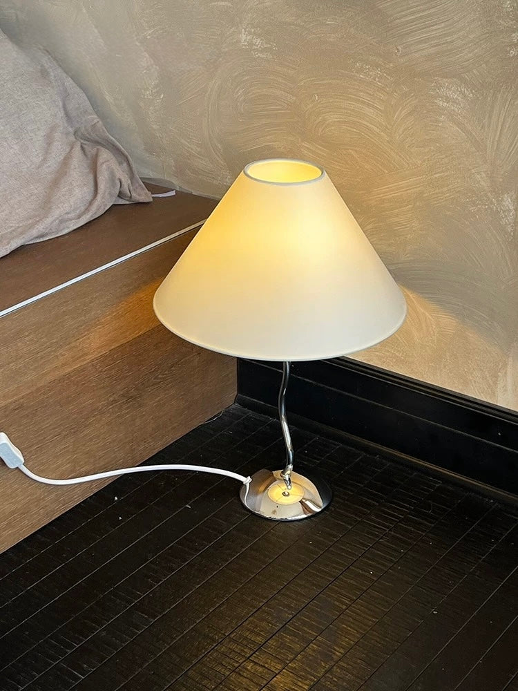 Small table lamp with a white shade on a dark surface next to a bed.