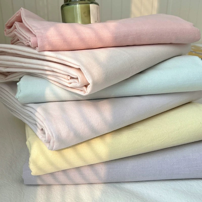 A stack of six folded cotton fabrics in pastel shades of pink, cream, mint, light purple, pale yellow, and lavender, arranged on a white surface with a small glass jar on top.