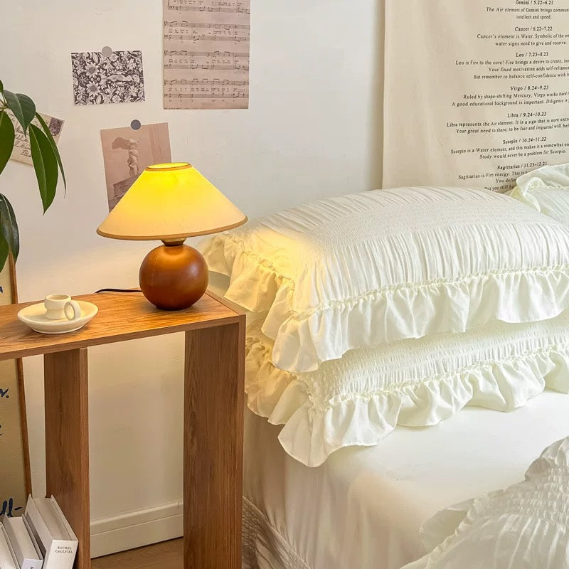Wooden side table with a lamp next to a bed with ruffled bedding.