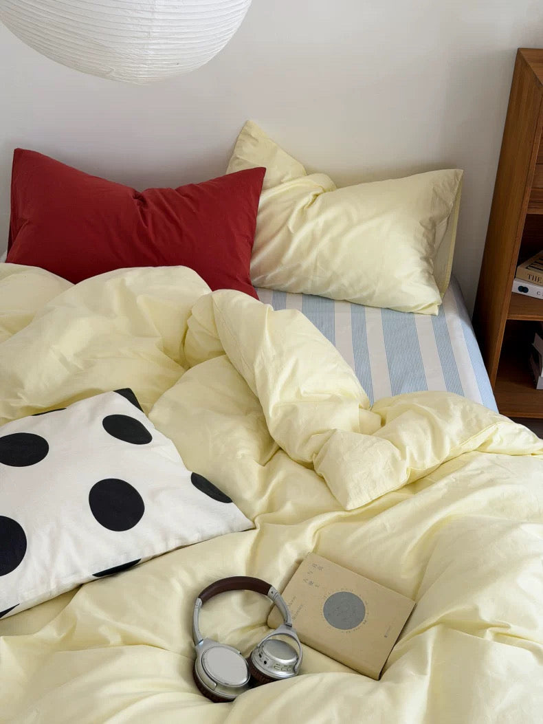A cozy bed styled with pale yellow bedding, a red pillow, a white pillow with black polka dots, blue and white striped sheets, over-ear headphones, and a small book in a bedroom setting.
