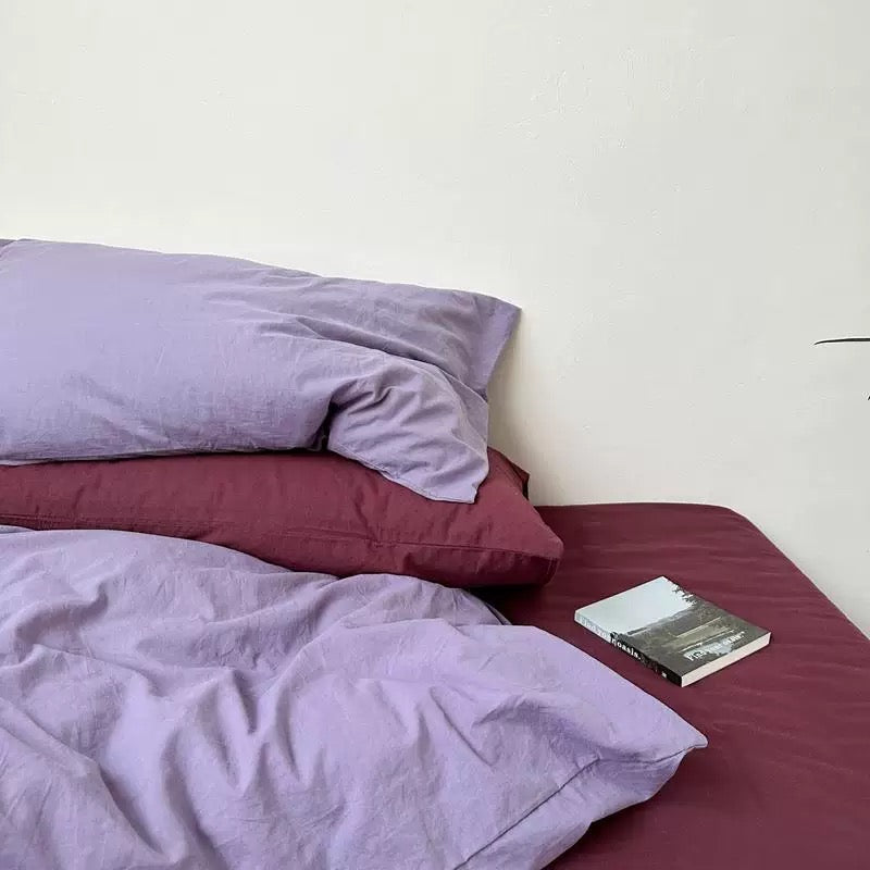 A lavender pillow cover rests above a plum pillowcase on burgundy bedding beside an open book.