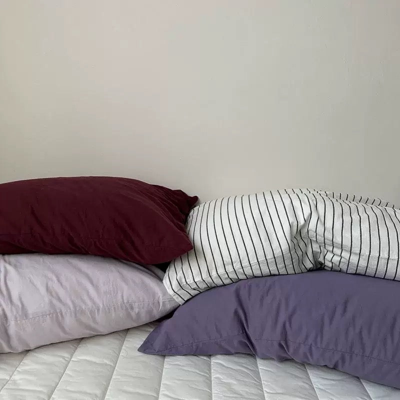 Lavender, plum, pale gray, and white striped pillowcases are arranged across a white bed.