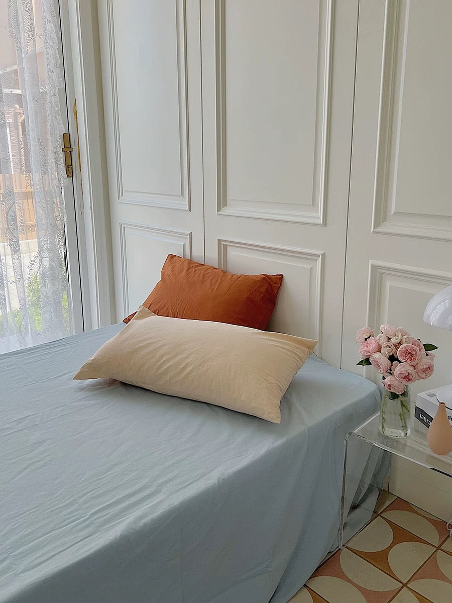 A light blue sheet drapes over a bed with an orange pillow behind a cream pillow.