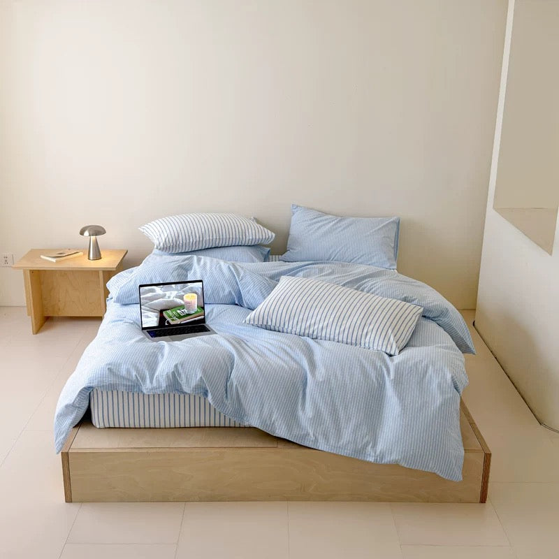 A light blue and white striped bedding set with matching pillows and duvet on a wooden platform bed, styled in a minimalist bedroom with a wooden nightstand and lamp.