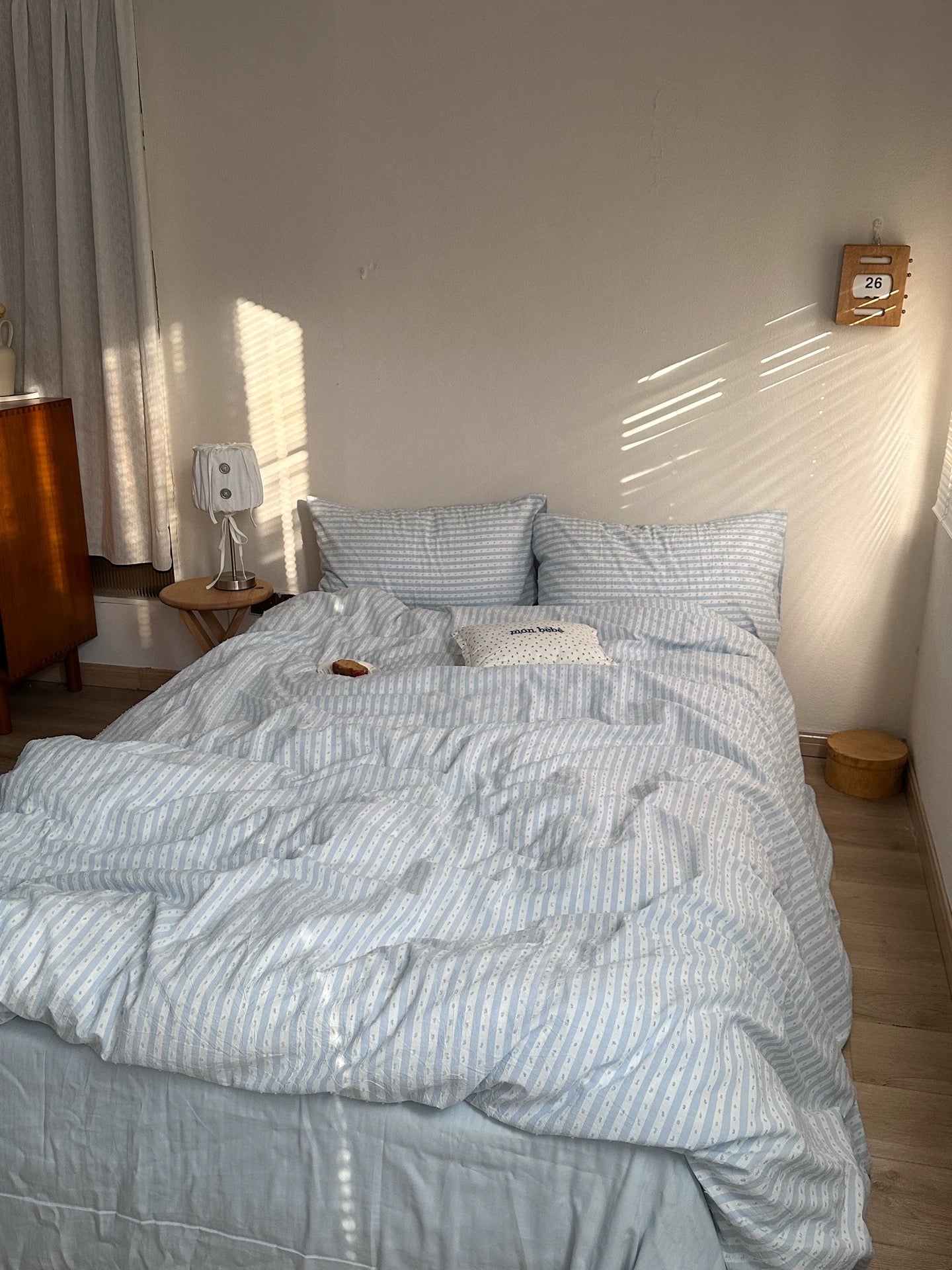 A bed dressed in light blue and white striped bedding, set in a softly lit bedroom with warm sunlight casting shadows on the wall.