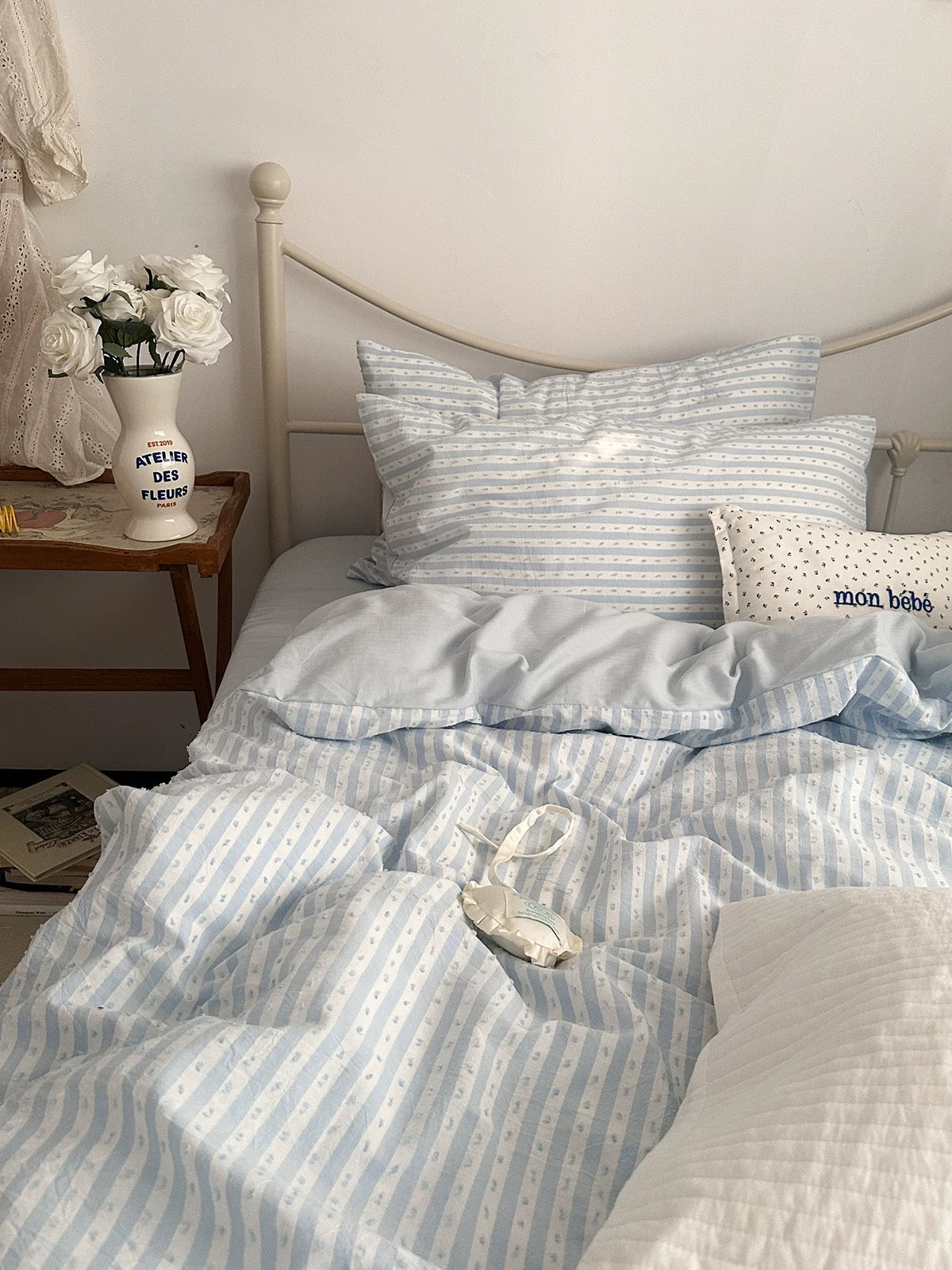 A neatly made bed in a bedroom setting featuring a light blue and white striped bedding set with matching pillows and duvet. A small wooden bedside table with a vase of white roses is visible on the left.