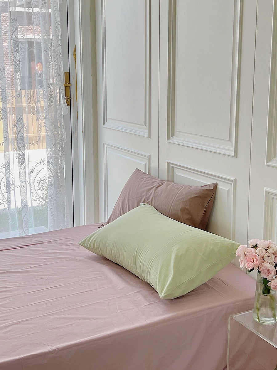 A light pink sheet covers the bed beneath a pale green pillow and a taupe pillow.