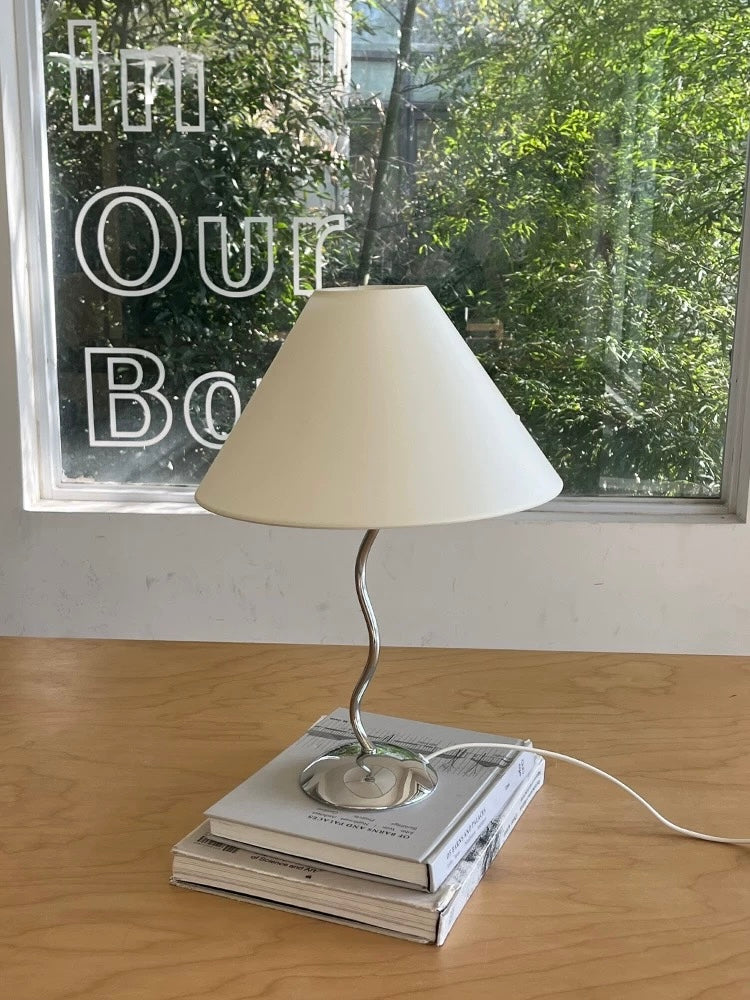 Table lamp with a white shade on top of books on a wooden surface, with a window in the background.