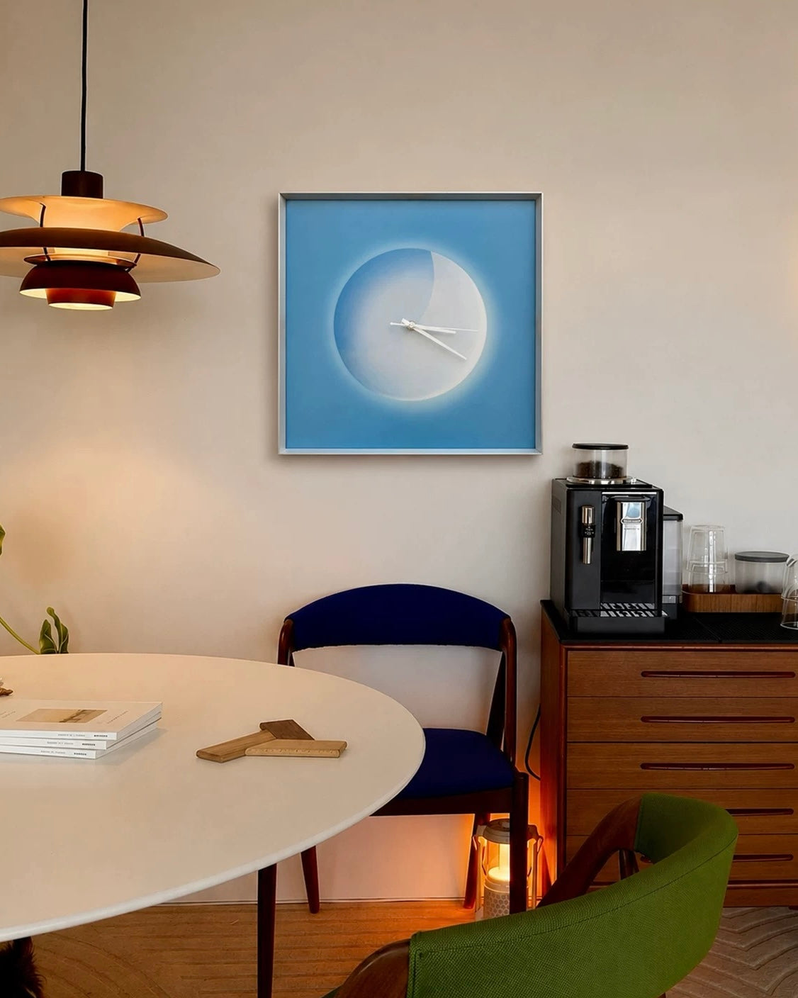 A square framed wall clock with a minimalist blue and white moon design hangs on a beige wall in a modern dining room setting.
