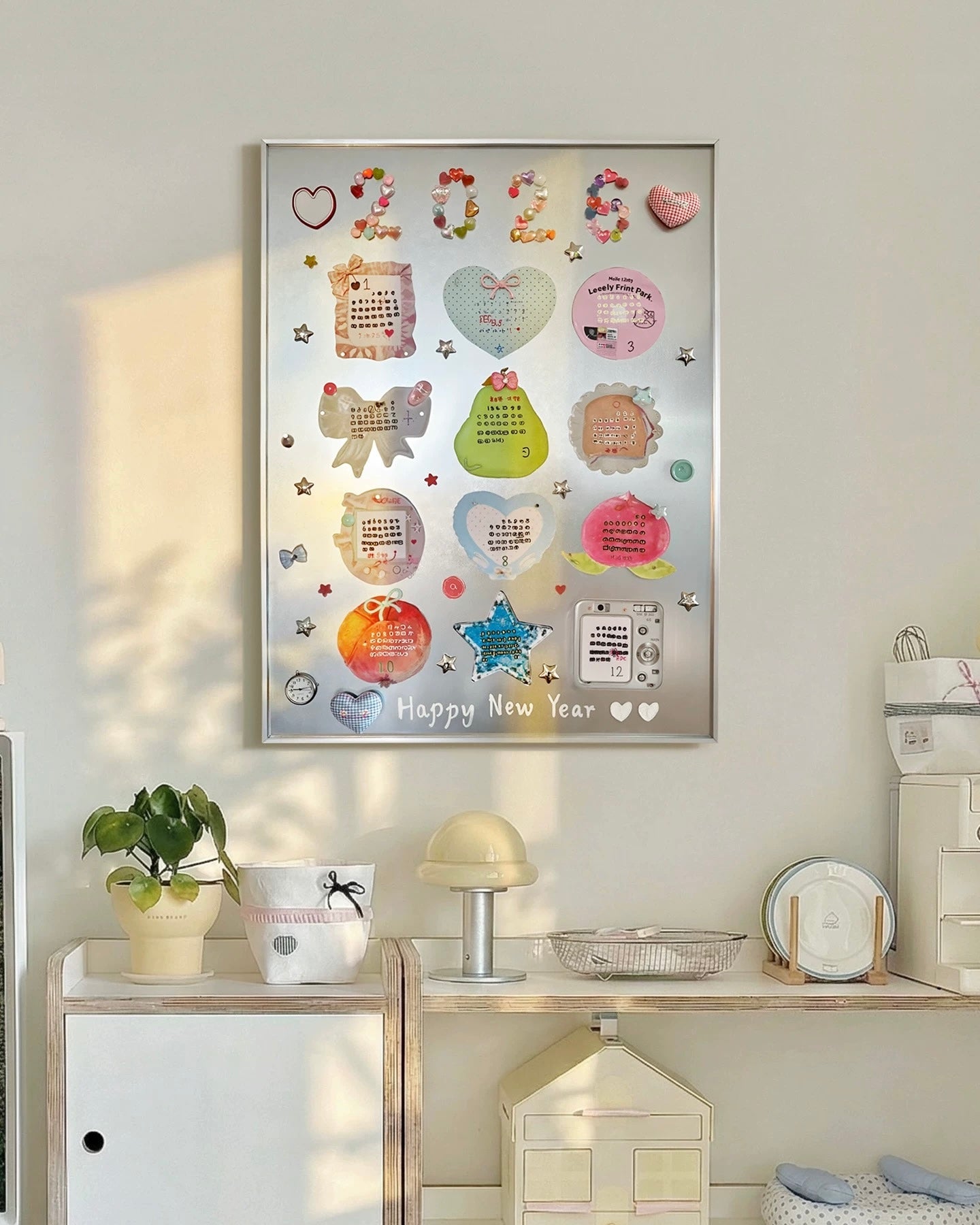 A framed New Year 2023 collage print with colorful stickers, hearts, stars, and text, displayed on a light beige wall above a styled shelf with a plant, lamp, and small house decor.