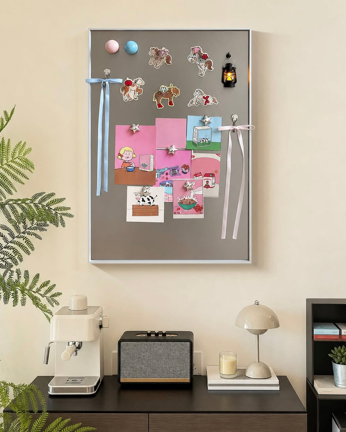 A gray magnetic refrigerator door board mounted on a beige wall, decorated with colorful magnets, ribbons, and pinned cards. Below the board, a dark wood console table holds a cream espresso machine, a small speaker, a lamp, and other decor.