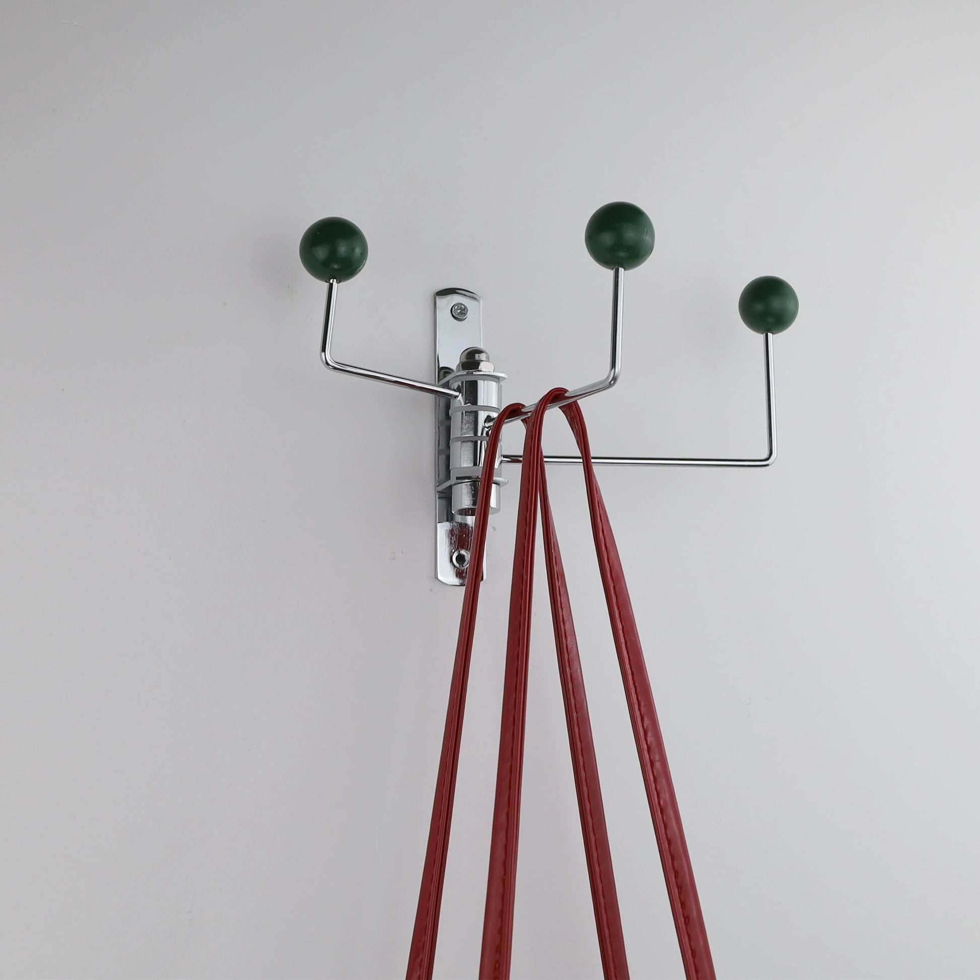 A wall-mounted three-hook coat rack with chrome metal arms and dark green spherical finials, shown against a plain light gray wall with red straps hanging from the center hook.