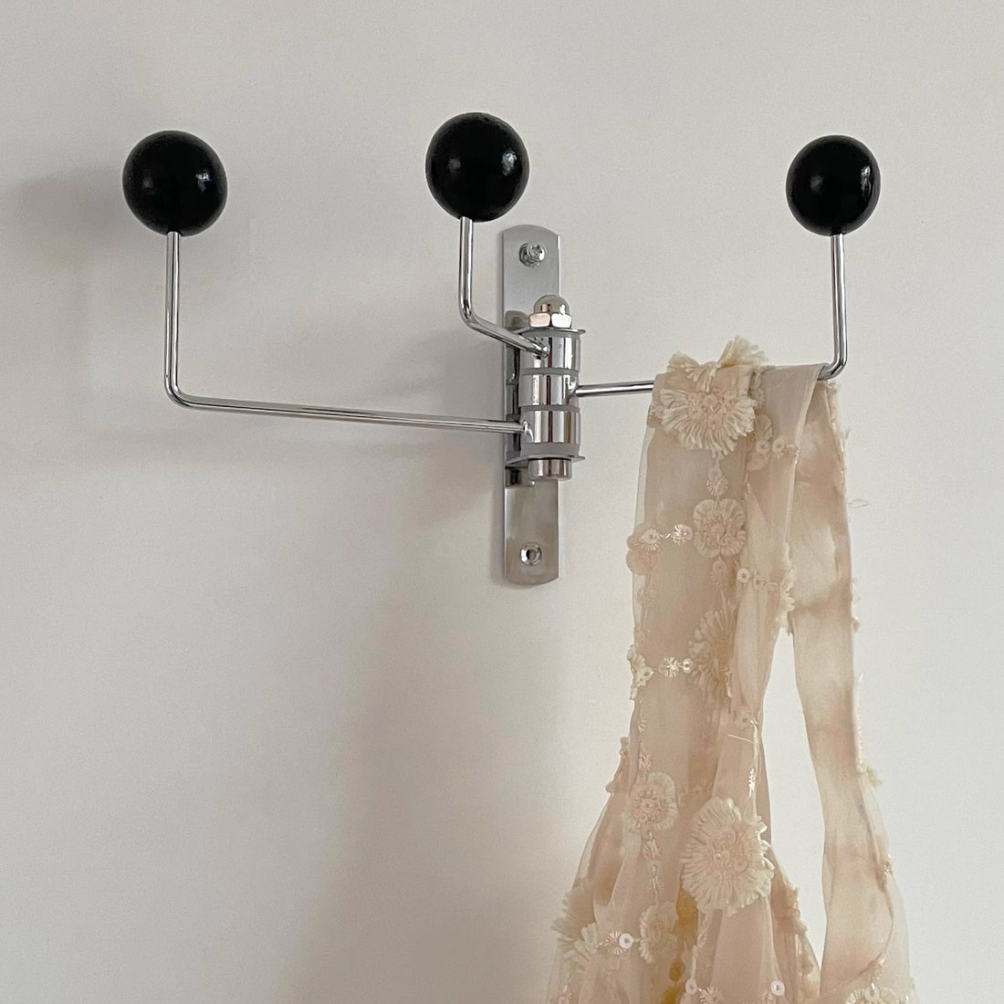 A cream woven tote bag with brown leather handles hangs from a silver wall hook with three cream spherical finials, against a plain off-white wall. A paper tag reading 'de luxe style' is attached to the bag.