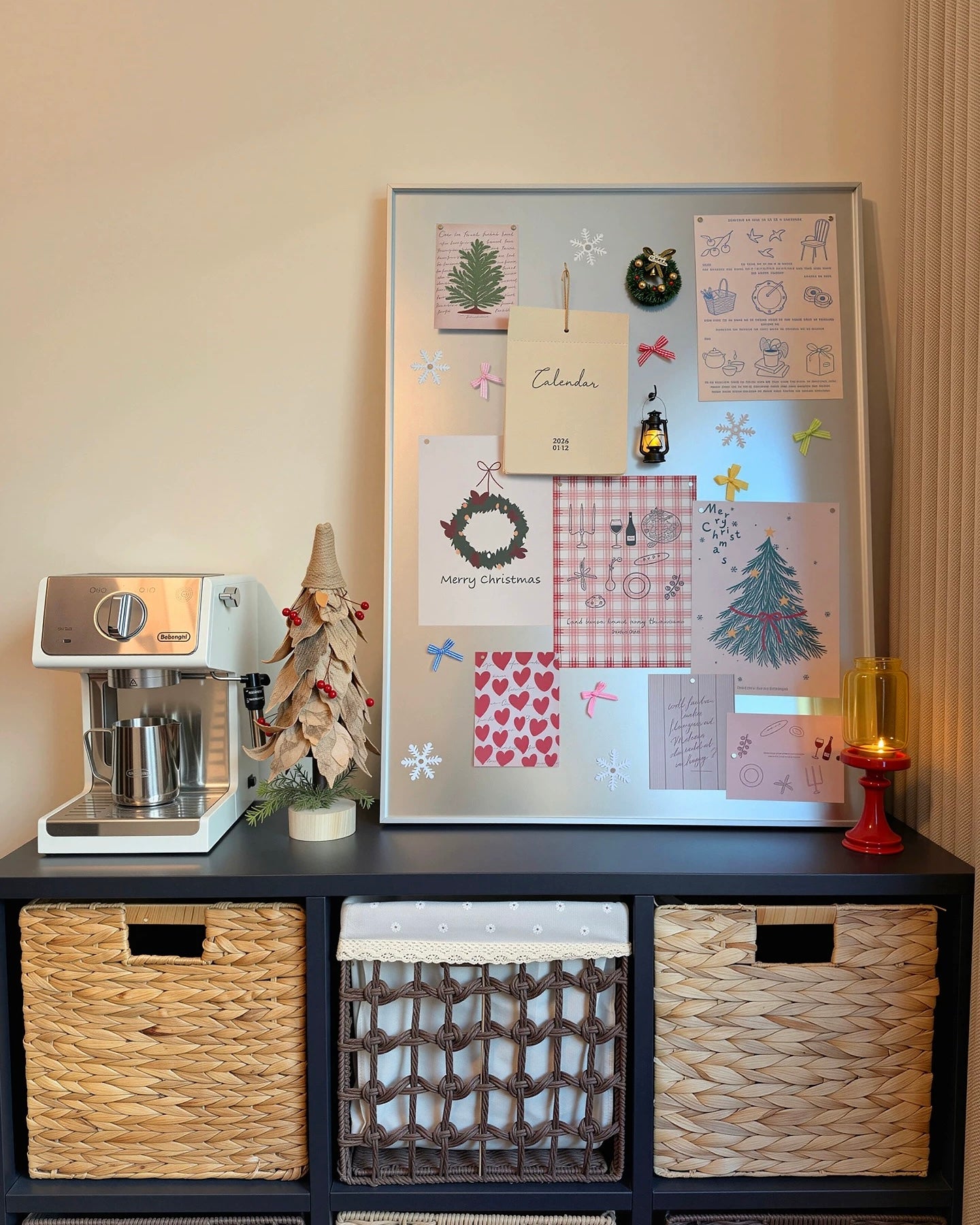 A styled interior scene showing a large white magnetic corkboard with holiday-themed cards, magnets, and illustrations, displayed on a black shelving unit with woven baskets. A white espresso machine sits on the left, and a lit red candle holder glows on the right.