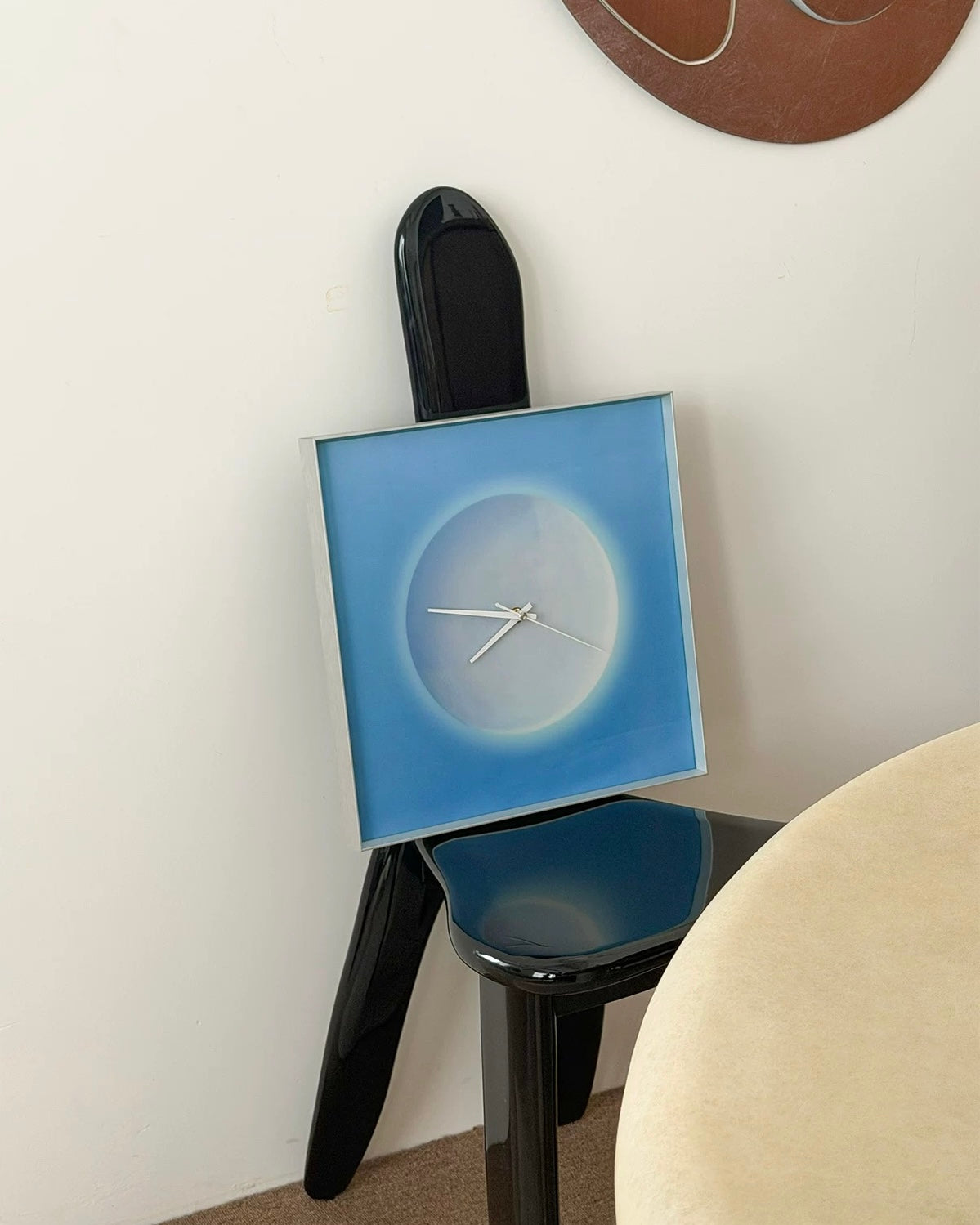 A square wall clock with a blue and white gradient design, leaning against a black chair in a room setting.