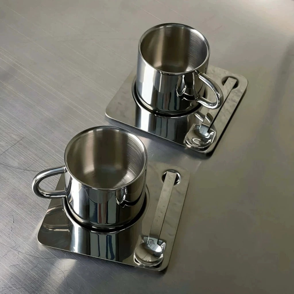 Two metal cups with handles on a matching metal tray on a reflective surface.