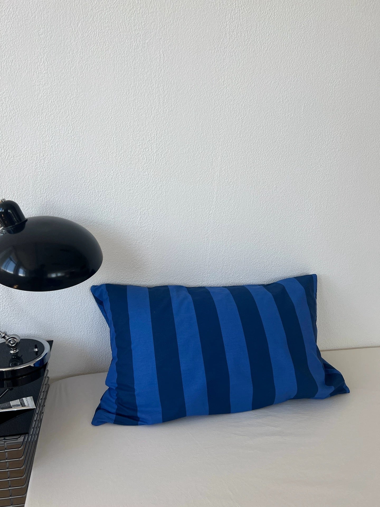 A rectangular throw pillow with vertical blue and navy stripes resting on a white bed against a white wall, with a black desk lamp visible on the left.