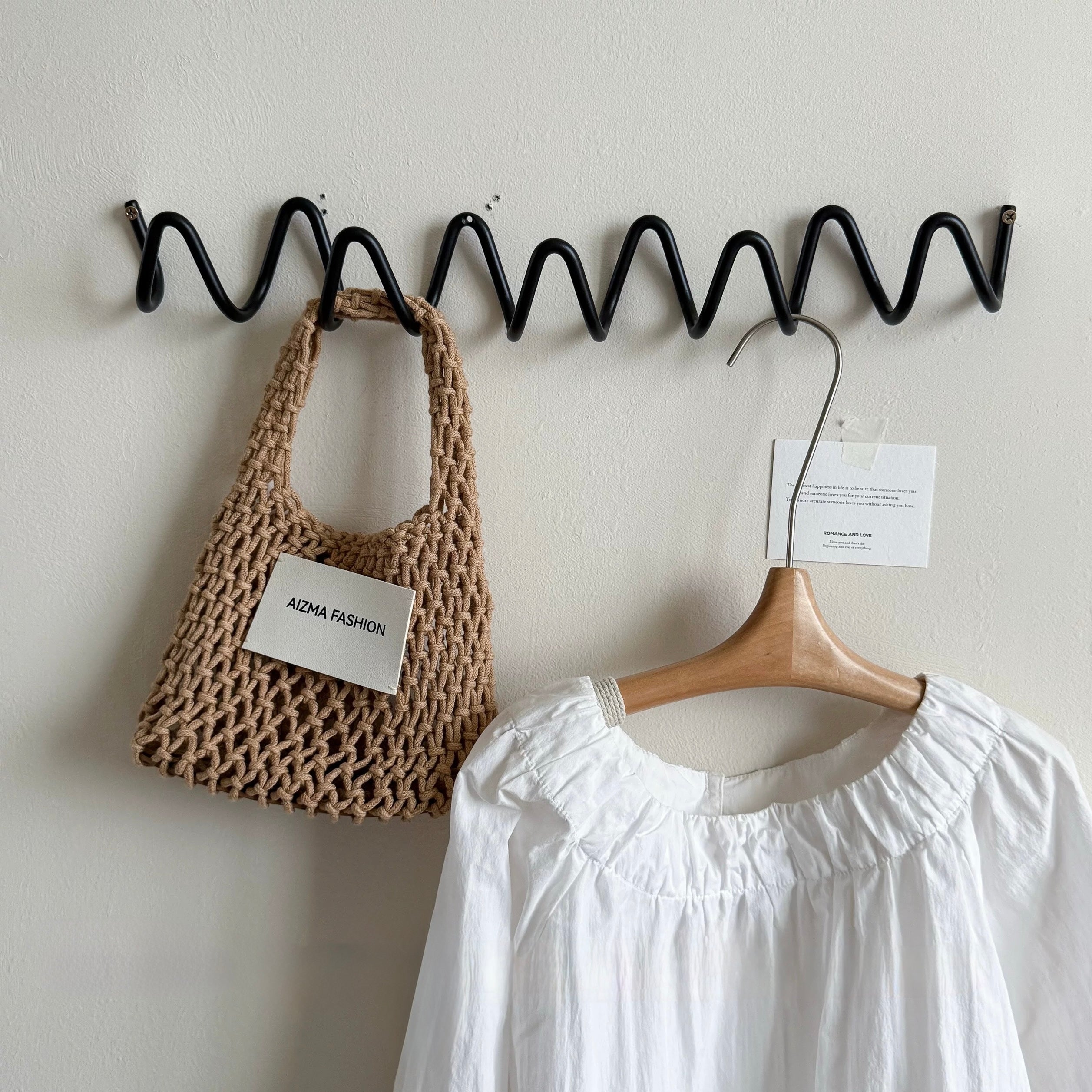 Woven bag and white garment on a black zigzag hook against a light gray wall.