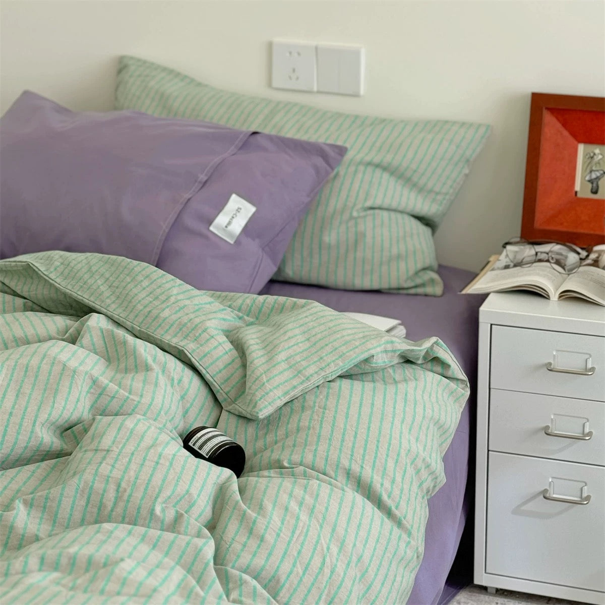 Bed with green and purple bedding, pillows, and a nightstand with an open book.