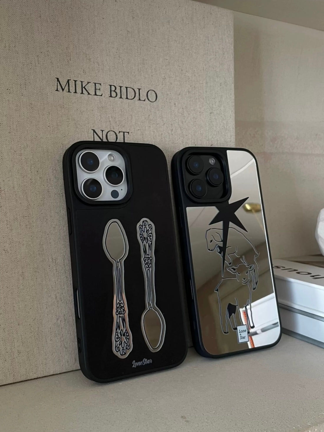 A mirror phone case in black with two vintage spoons stands beside the silver lamb and pointed star design.
