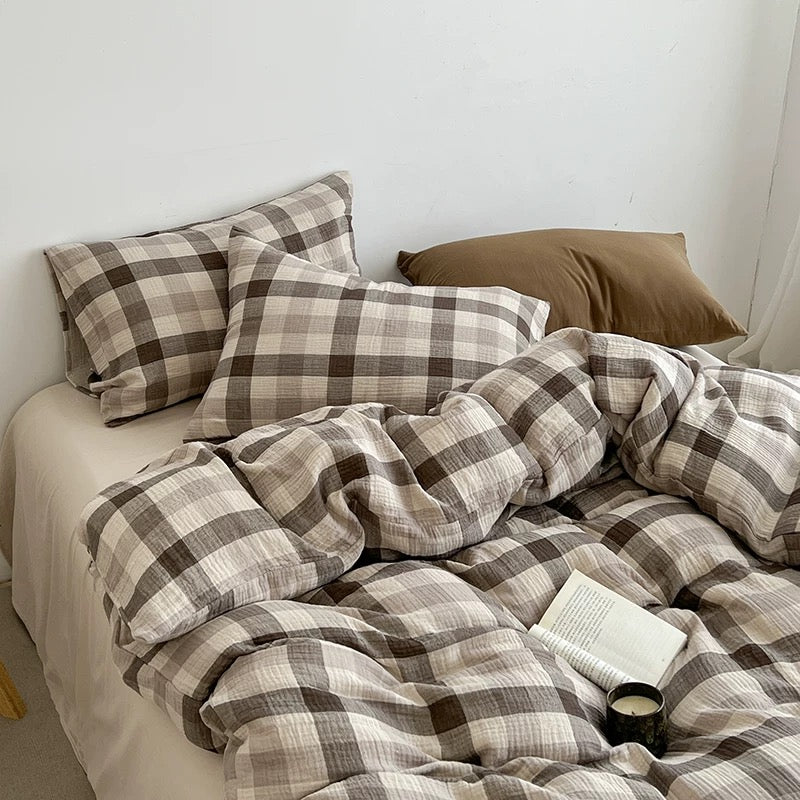 A bed styled with a brown and beige plaid bedding set, including a duvet cover and matching pillowcases, with a solid brown pillow and a white wall in the background.