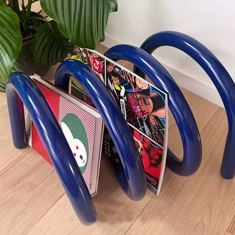 Blue metal magazine rack holding colorful books on a wooden floor with a plant in the background.