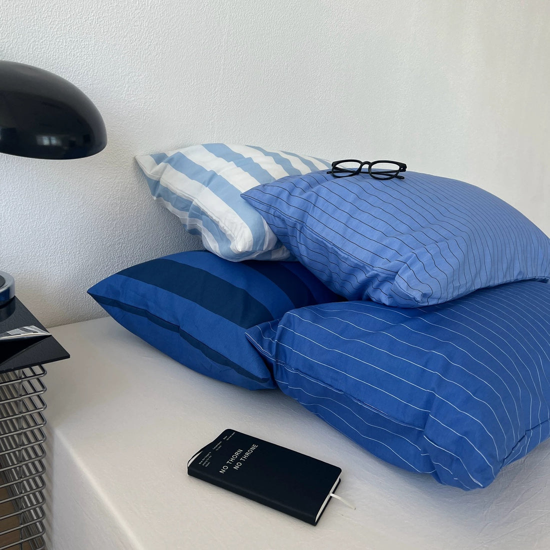 Three blue and white striped and pinstriped pillows arranged on a bed next to a black desk lamp and notebook.
