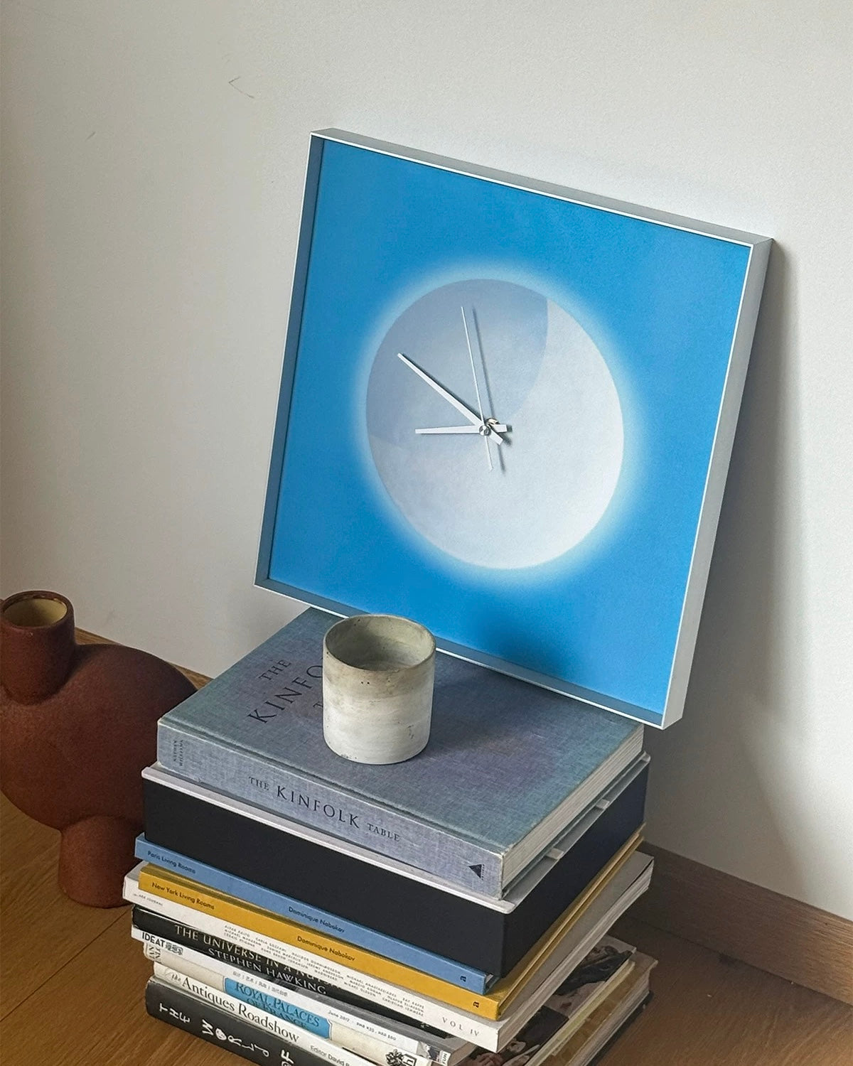 A square wall clock with a blue and white moon design, leaning on a stack of books next to a small ceramic cup against a plain wall.