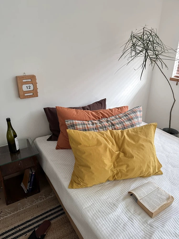 A mustard yellow pillowcase sits at the front of a bed with plaid and brown designs behind it.