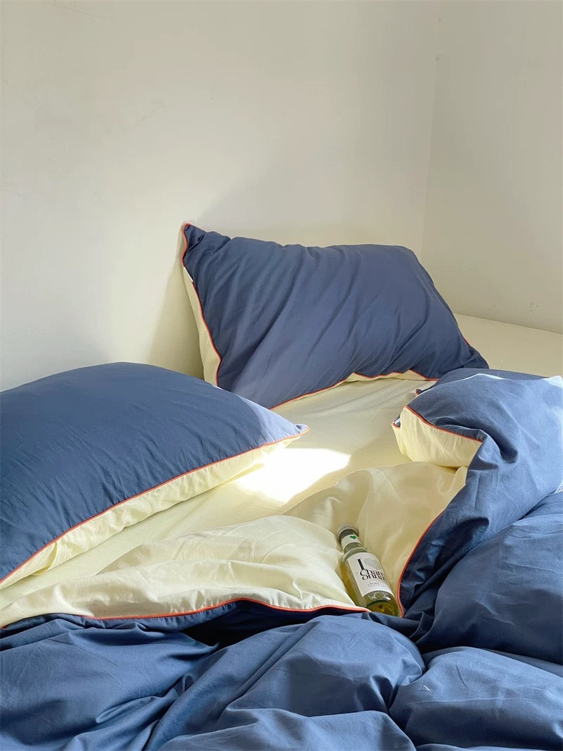 Unmade bed with blue bedding and a bottle of beer on a white wall background