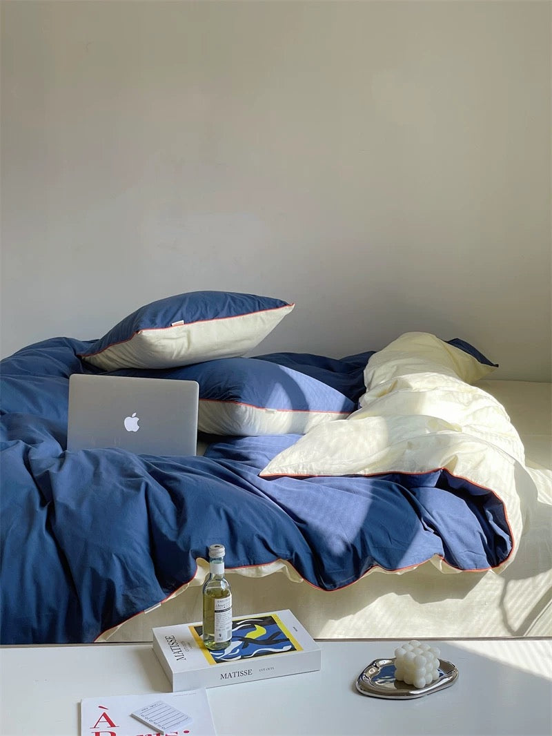 Bed with blue bedding and white pillows, laptop, and small table with items on a light wooden floor.
