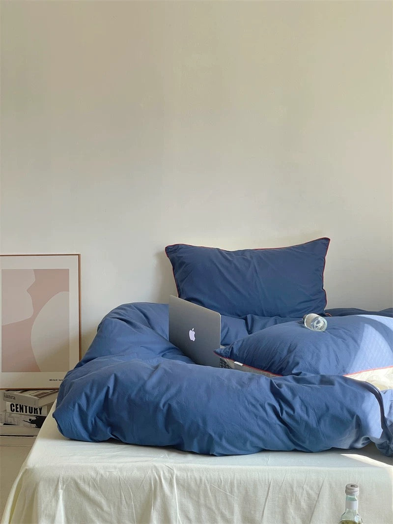 Blue comforter on a bed with a laptop and water bottle, against a plain wall.