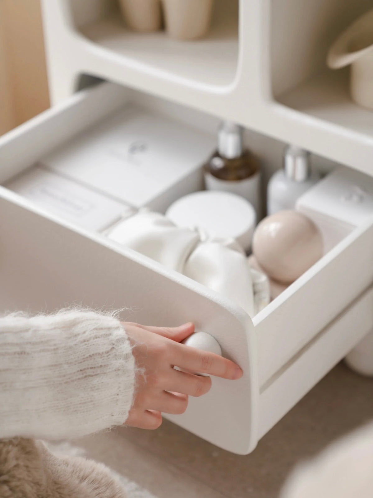 An open white nightstand drawer is shown close up, with a hand on the round knob beneath the shelves.