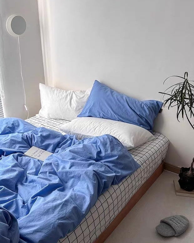 Bed with blue bedding and pillows in a room with a plant and slippers on the floor.