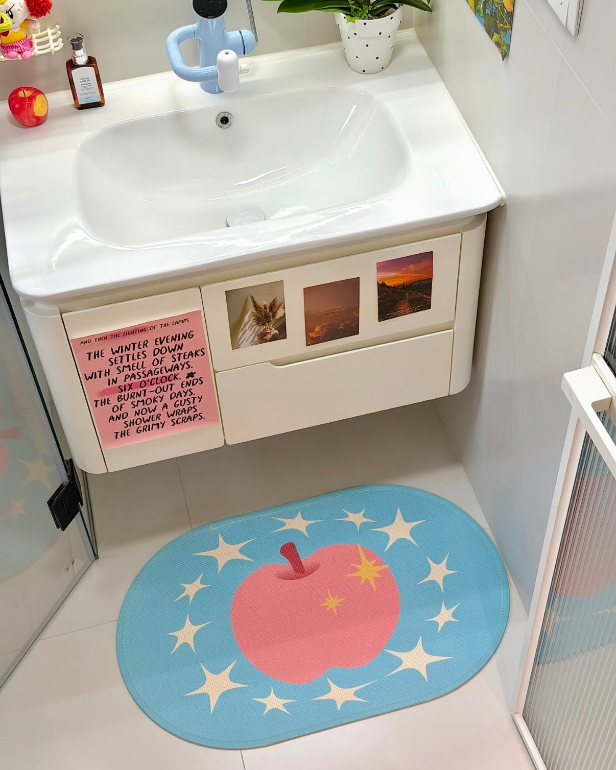 A bathroom floor mat with a pink apple and white stars on a light blue background, placed in front of a white vanity cabinet.
