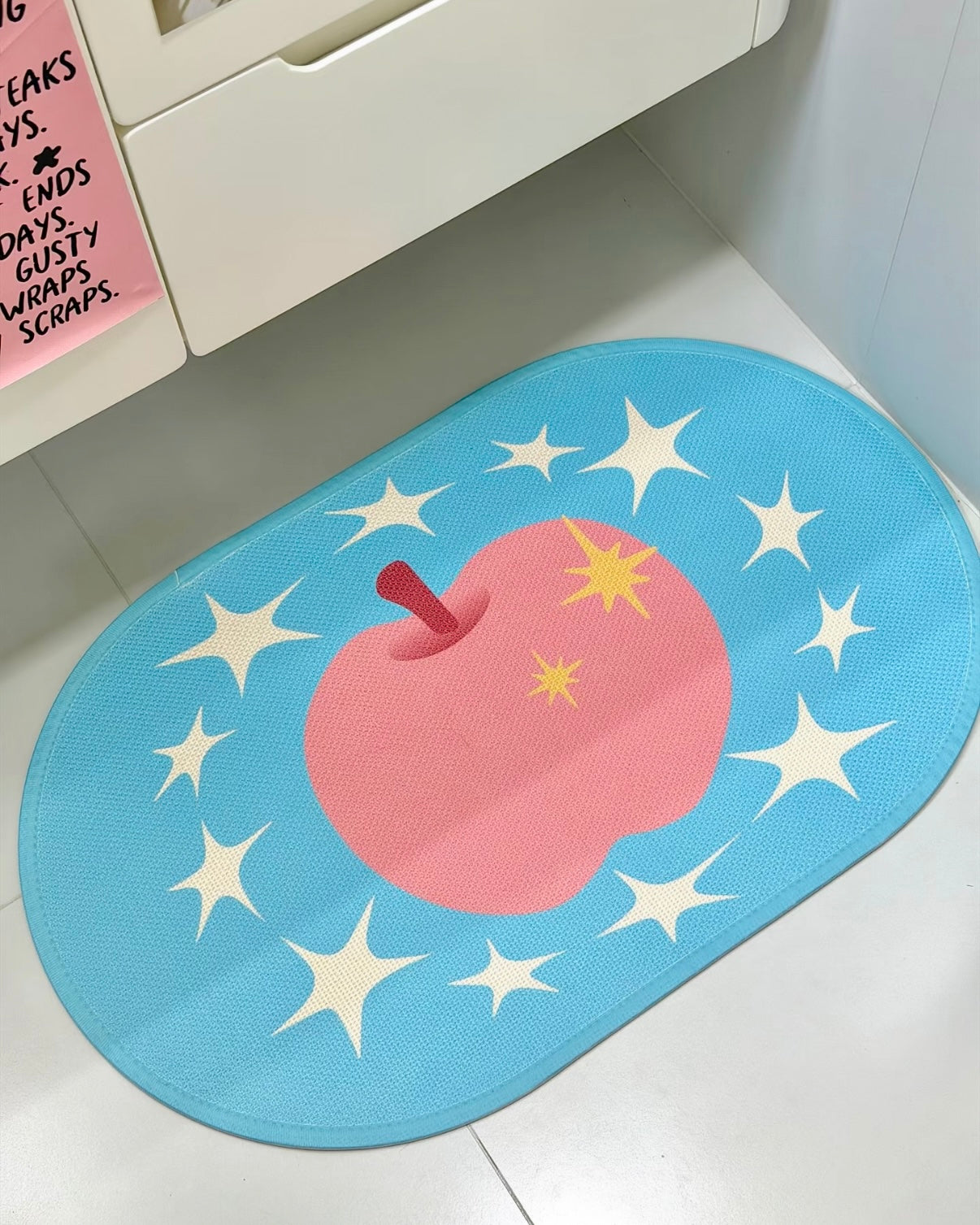 An oval blue floor mat featuring a large pink apple with a red stem and yellow stars, surrounded by white starburst shapes, placed on a white tiled floor next to a white cabinet.
