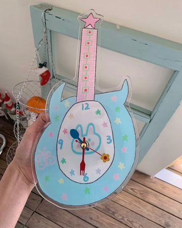 A hand holds a hand-painted acrylic wall clock shaped like a cat face with a guitar neck. The clock features a light blue cat face, a pink plaid neck, and colorful star and number details, set against a rustic indoor background.