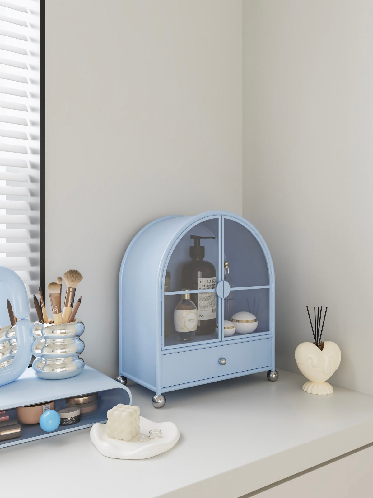 A blue pastel makeup organizer shown closer has an arched top, glass doors and a silver drawer knob.