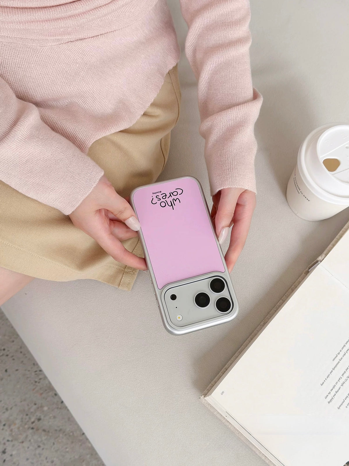 Pastel pink phone case held at an angle, showing a pale back, a silver colored border and black lettering.