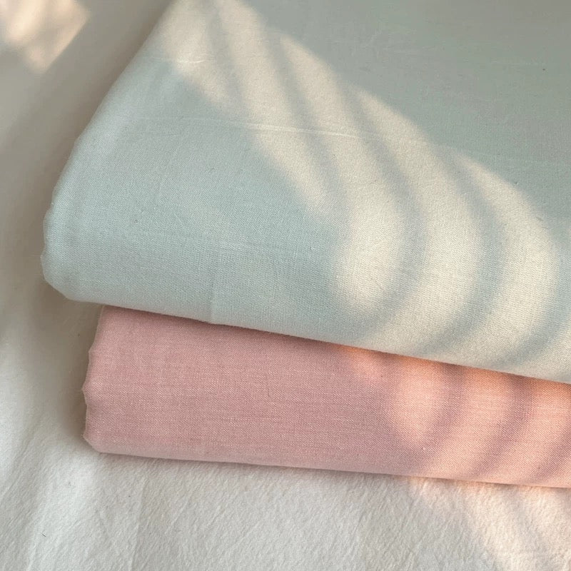 Two folded cotton fabrics, one in pale mint green and one in soft blush pink, stacked on a white textured surface with dappled sunlight and leaf shadows.