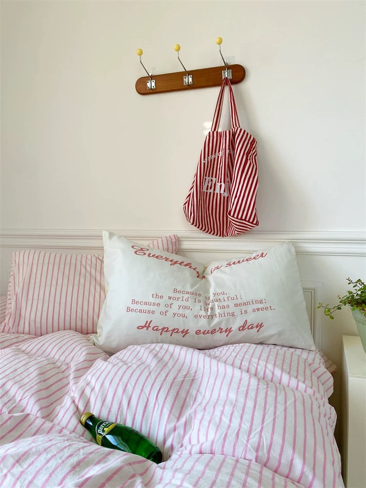 Bed with pink striped bedding and a pillow with text, next to a red and white bag hanging on a hook.