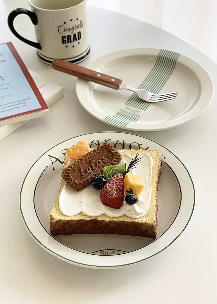 A styled tabletop scene with a white ceramic plate printed with the word 'Asparagus' in green, holding a small dessert with berries and cream, alongside a fork. A white mug with a black handle and 'Mine' printed on it sits nearby, next to stacked books including one titled 'What I Wish I Knew When I Was 20' by Tina Seibel.