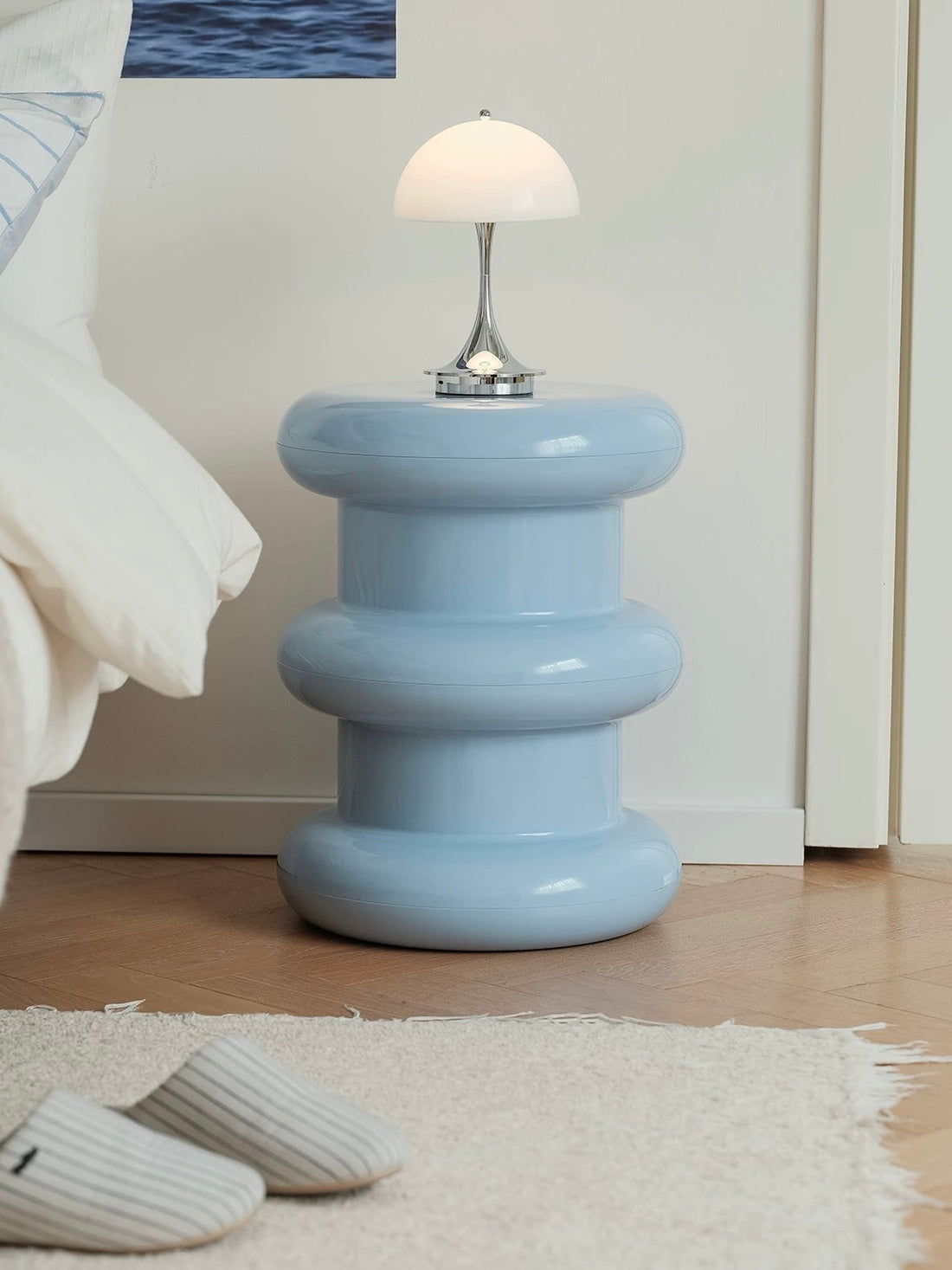A light blue, tiered, screwdriver-shaped side table with a glossy finish, styled with a magazine on top in a bright room with light wood floors and white furniture.