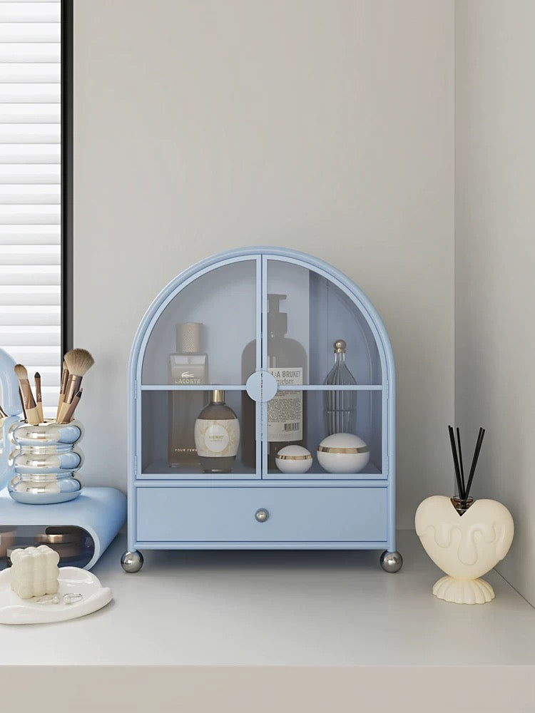 A blue perfume cabinet has arched glass doors, silver ball feet and a lower drawer beneath the bottles.