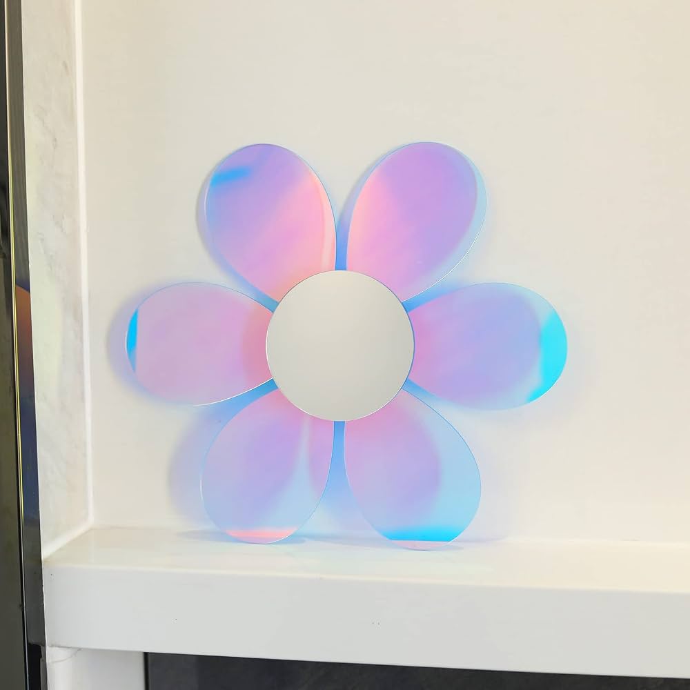 A flower-shaped wall mirror with iridescent petals reflecting pink, orange, and blue light, mounted on a white wall above a gray wire shelf with a yellow decorative object.
