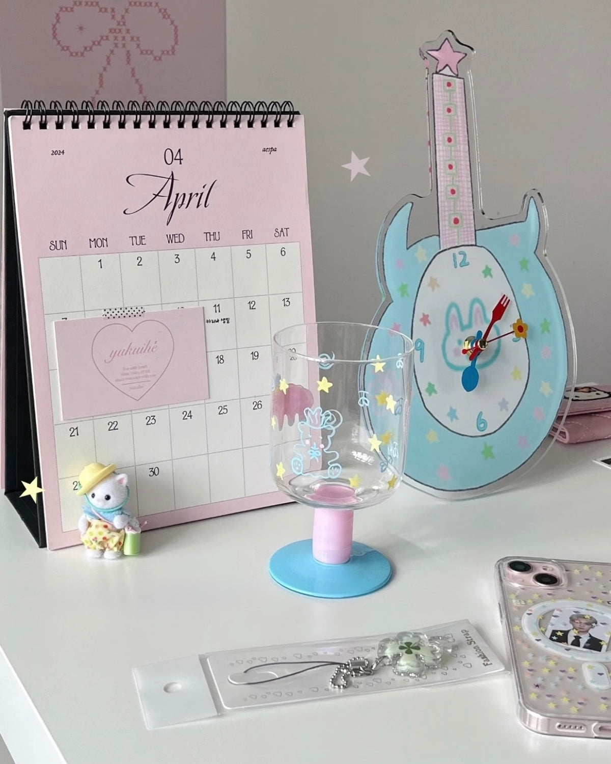 A styled desk scene with a pink spiral calendar showing April, a light blue guitar-shaped clock, a clear glass candle holder with a pink base and star/yellow accents, a small plush figure, and a phone case on a white surface.