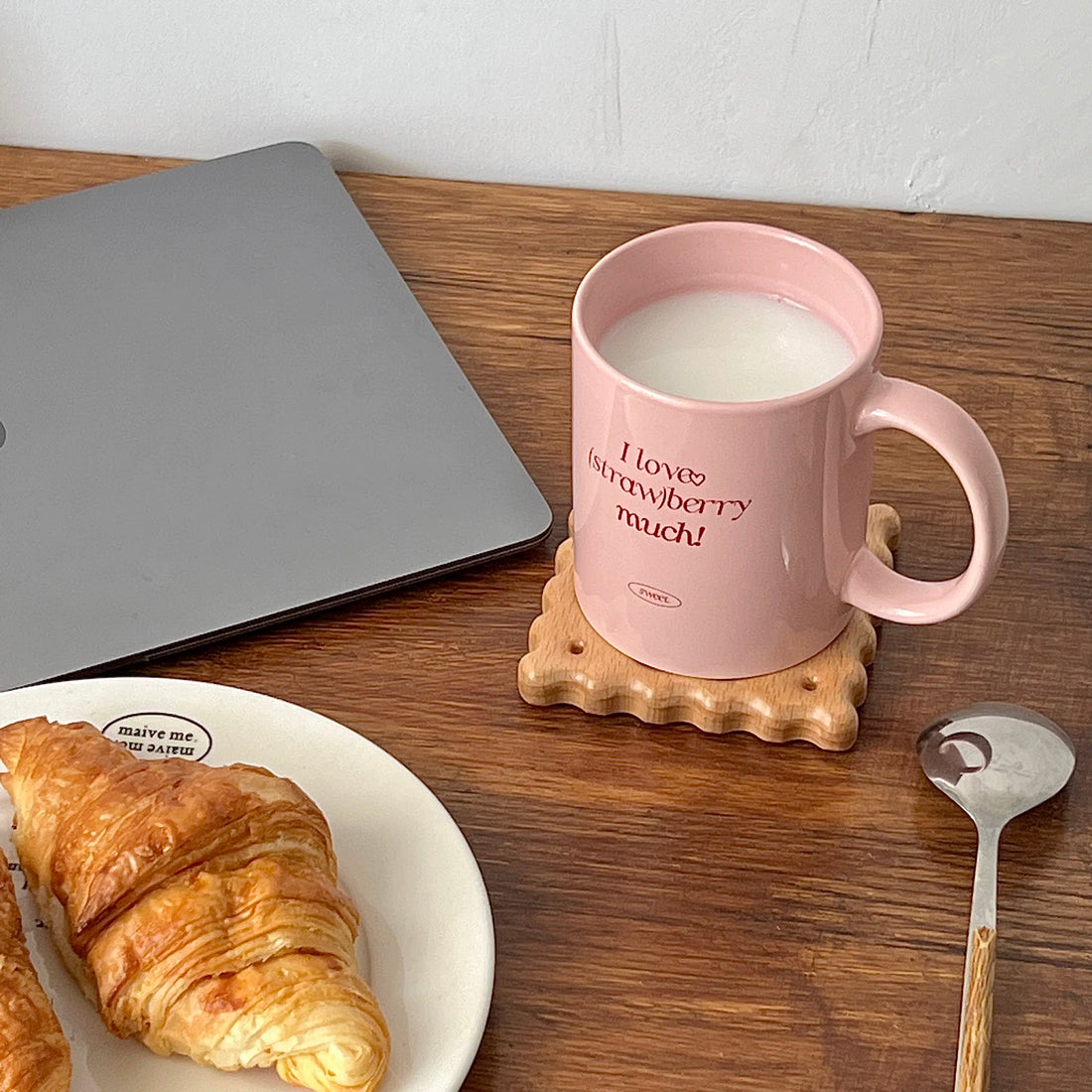 A pink ceramic mug filled with milk sits on a wooden table next to a plate with a croissant. The mug has the text 'I love strawberry much!' printed in red, with a small oval logo below that reads 'SWEET'.