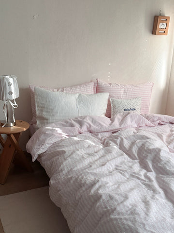 A bed dressed in light pink and white striped bedding, including a duvet cover, matching pillowcases, and several pillows, set in a bedroom with a wooden side table and a small wall clock.